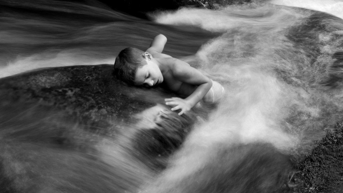 Un enfant allongé sur un rocher au milieu du courant d'un petit ruisseau ardéchois. Photographie en noir et blanc de Matthieu Dupont