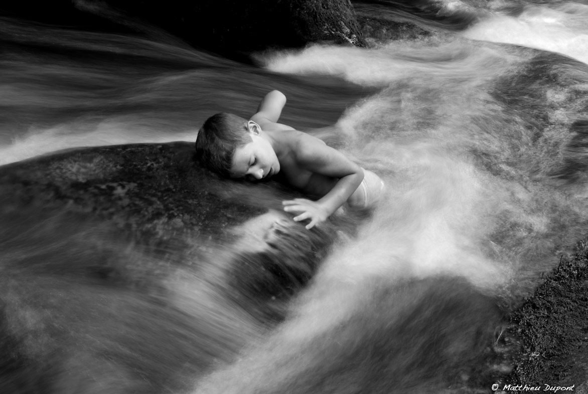 Un enfant allongé sur un rocher au milieu du courant d'un petit ruisseau ardéchois. Photographie en noir et blanc de Matthieu Dupont